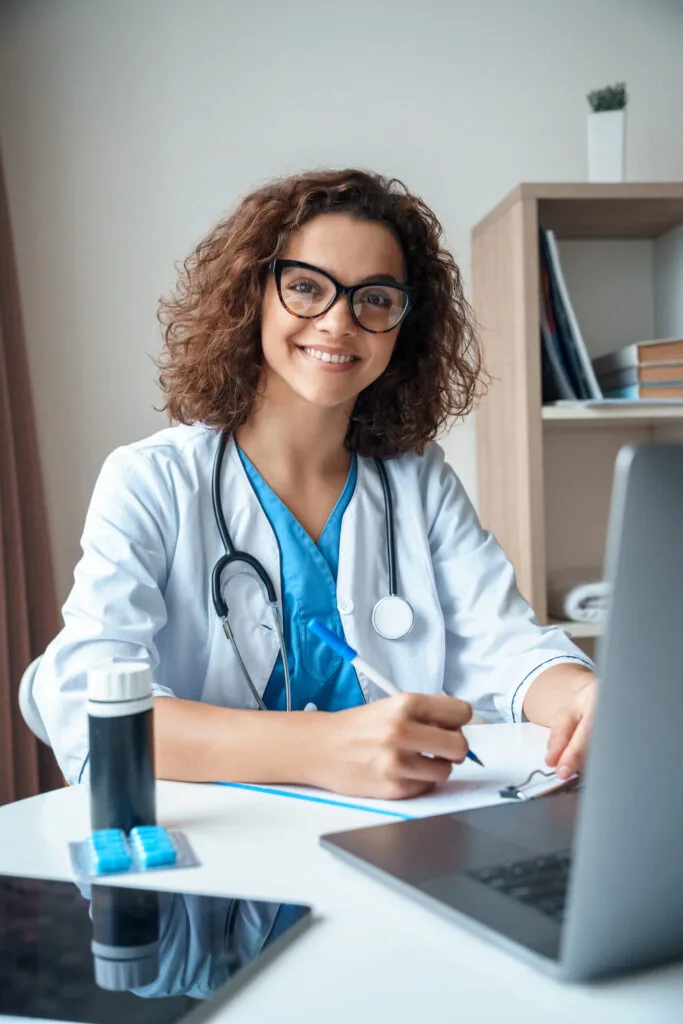 Vertical,Portrait,Of,Female,Doctor,Sitting,On,Work,Desk,And