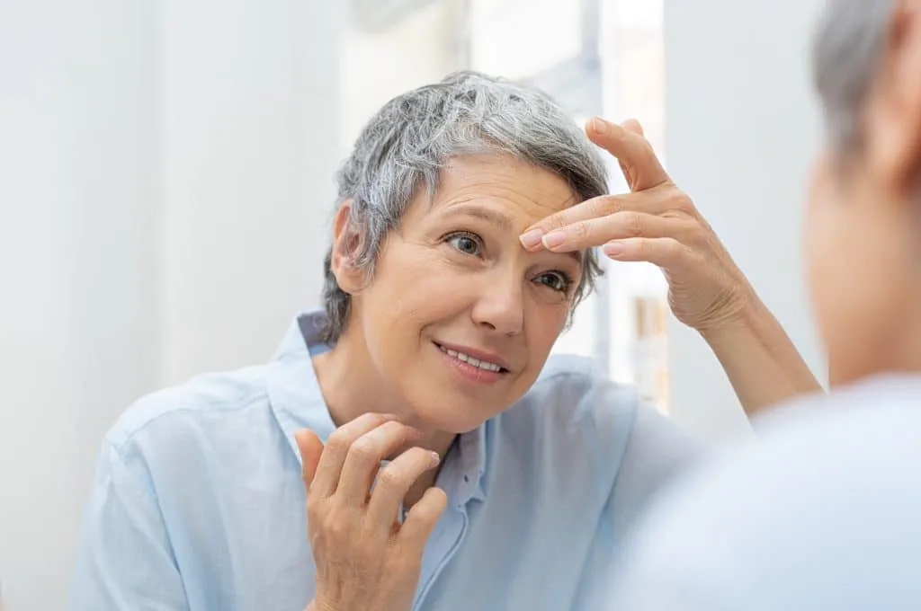An older woman examining the skin around her eye in a mirror