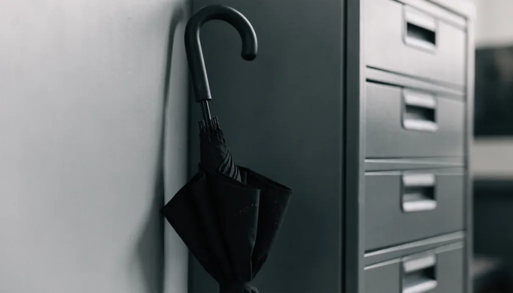 A closed umbrella resting against a plain office wall beside a filing cabinet, muted daylight