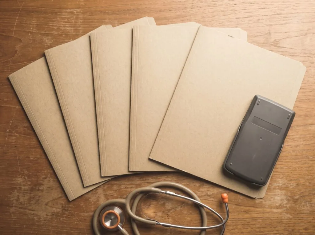 Six blank manila folders fanned out on a wooden desk beside a face-down calculator and a stethoscope